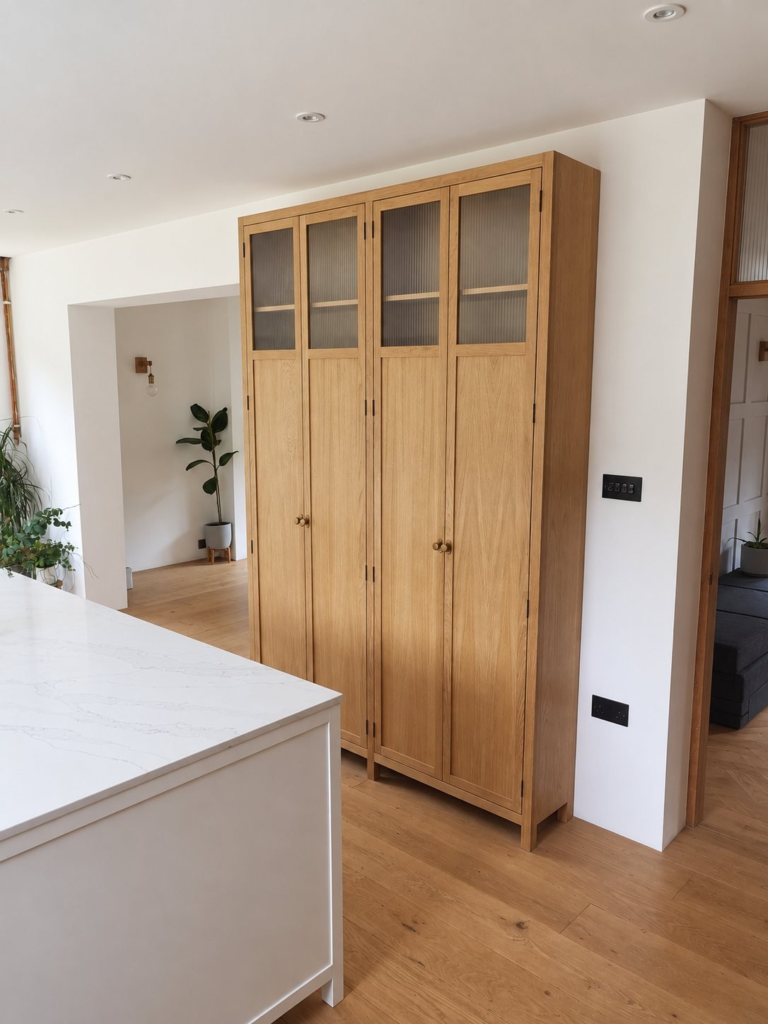 Tall oak larder cupboard with reeded glass upper doors and brass knobs, beside a white kitchen island